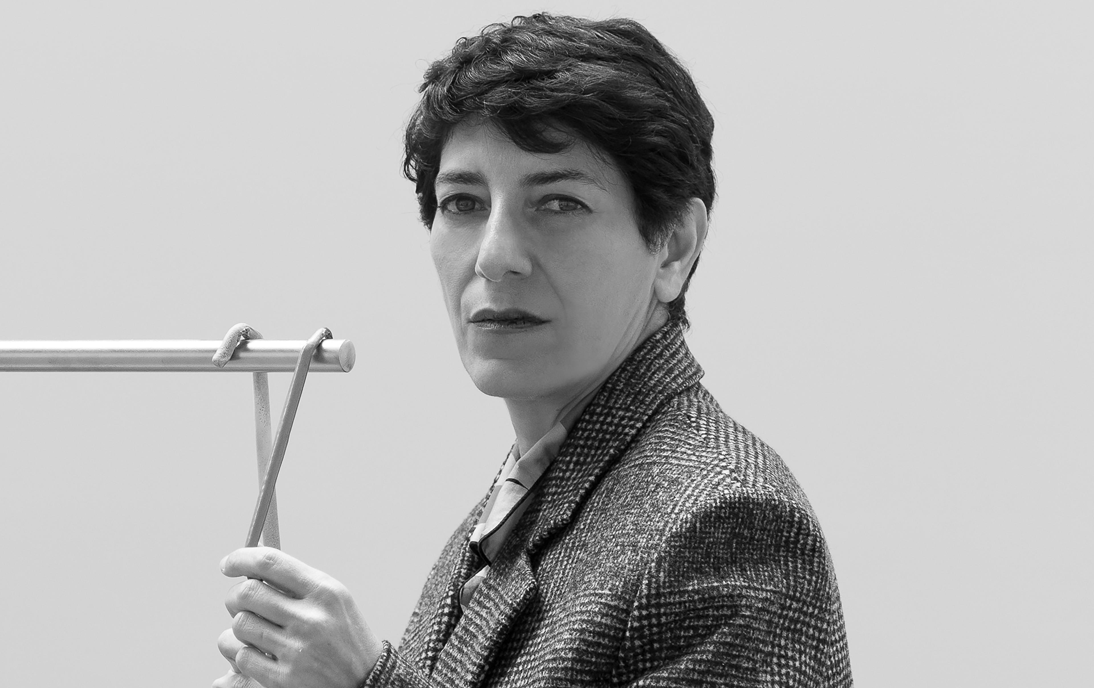 A black and white photo of Nairy Baghramian, a middle-aged woman with short dark cropped hair, wearing a tweed blazer and holding a wooden object in her hands. 