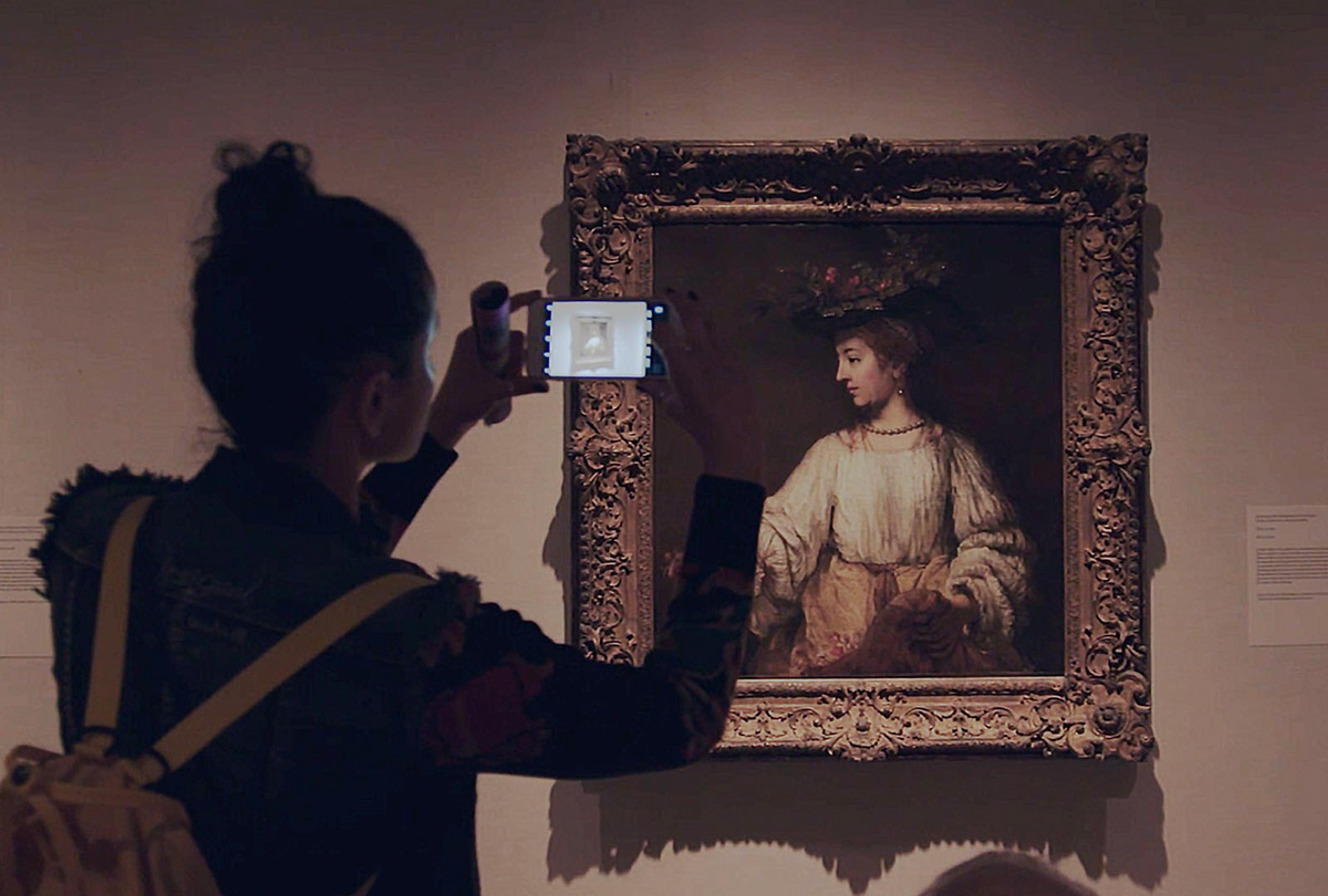 A woman holds up her phone to take a photo of a painting.