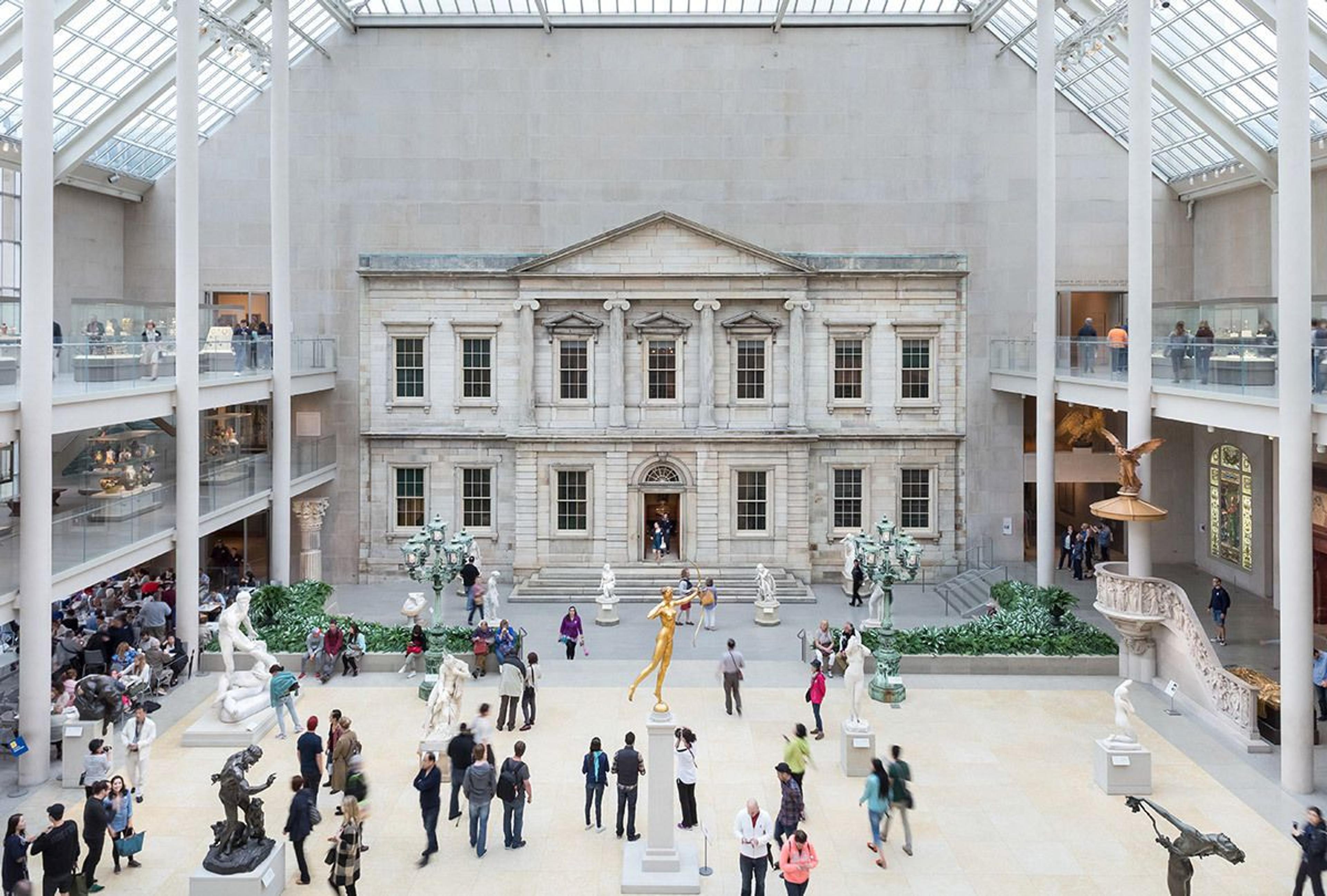 Visitors walking around an indoor courtyard with sculptures, a facade of a building is in the background.