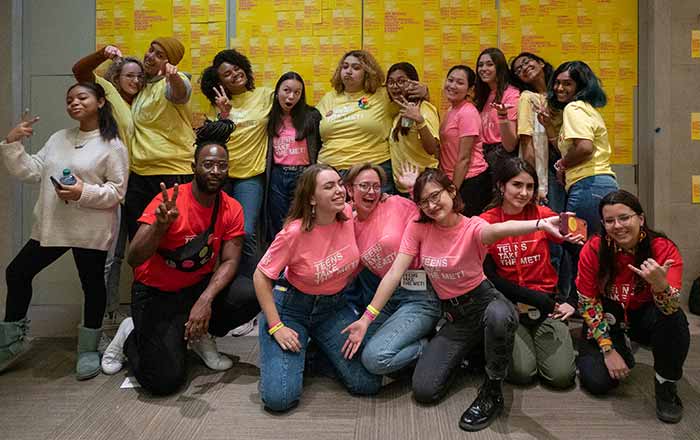 A group of teens are waving and making fun faces to the camera. They are wearing pink and yellow t-shirts, and look very happy.
