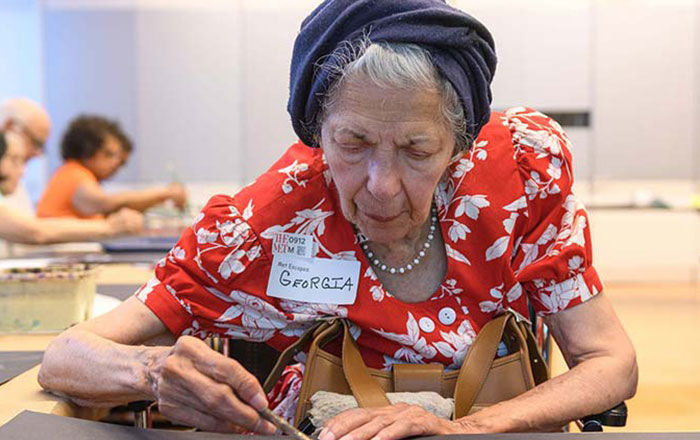 An elderly woman wearing the name tag "Georgia" works diligently on a drawing in a classroom of other participants
