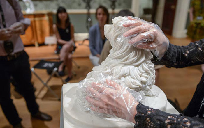 Two hands wearing clear gloves touch a small limestone sculpture of a curly-haired dog among a group of other seated participants in a gallery setting.