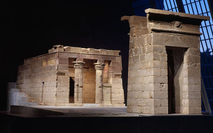 An ancient Egyptian sandstone temple in two separate parts in a large glass-enclosed gallery at dusk; the first part of the temple is a large squared arch, the second is a small rectangular building fronted by two columns topped with stylized leaves and plants