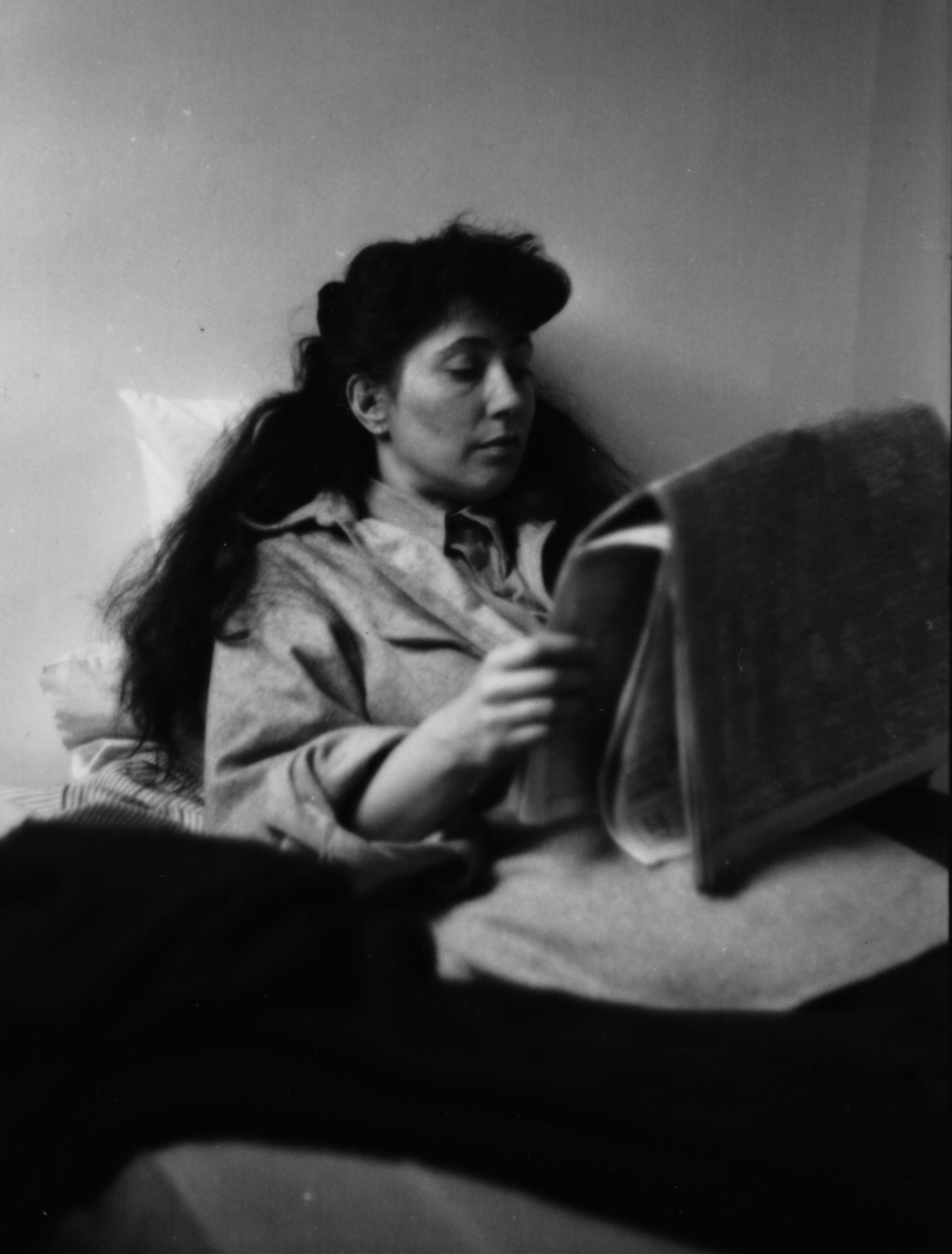 A black and white photograph of a young woman with long hair reading a newspaper in bed.