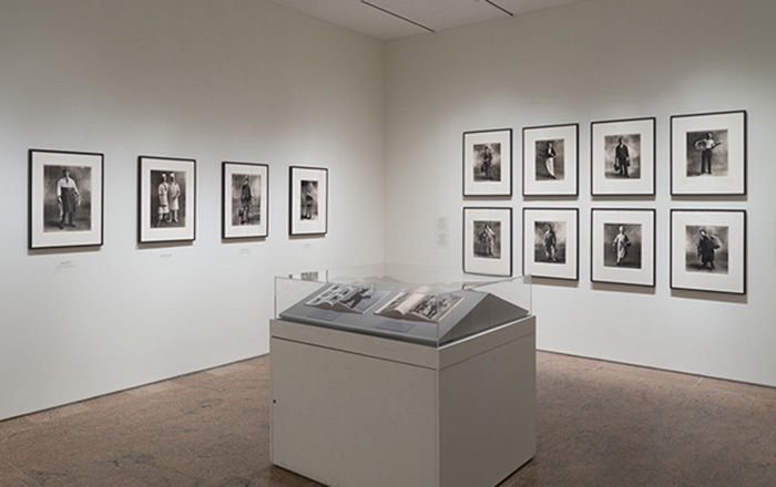 Photographs displayed in square frames hang on a white wall. More photography is displayed inside a glass case in the center of the room.