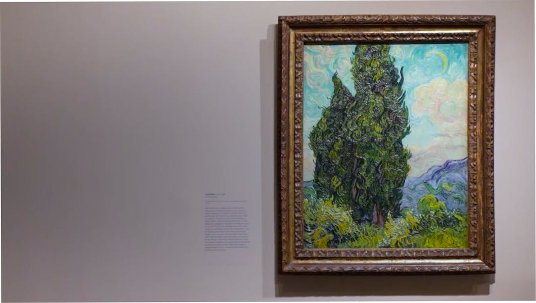 Vincent  Van Gogh’s Cypresses hangs on a wall in a wooden frame. The painting depicts tall trees on the left side with a swirling sky behind.